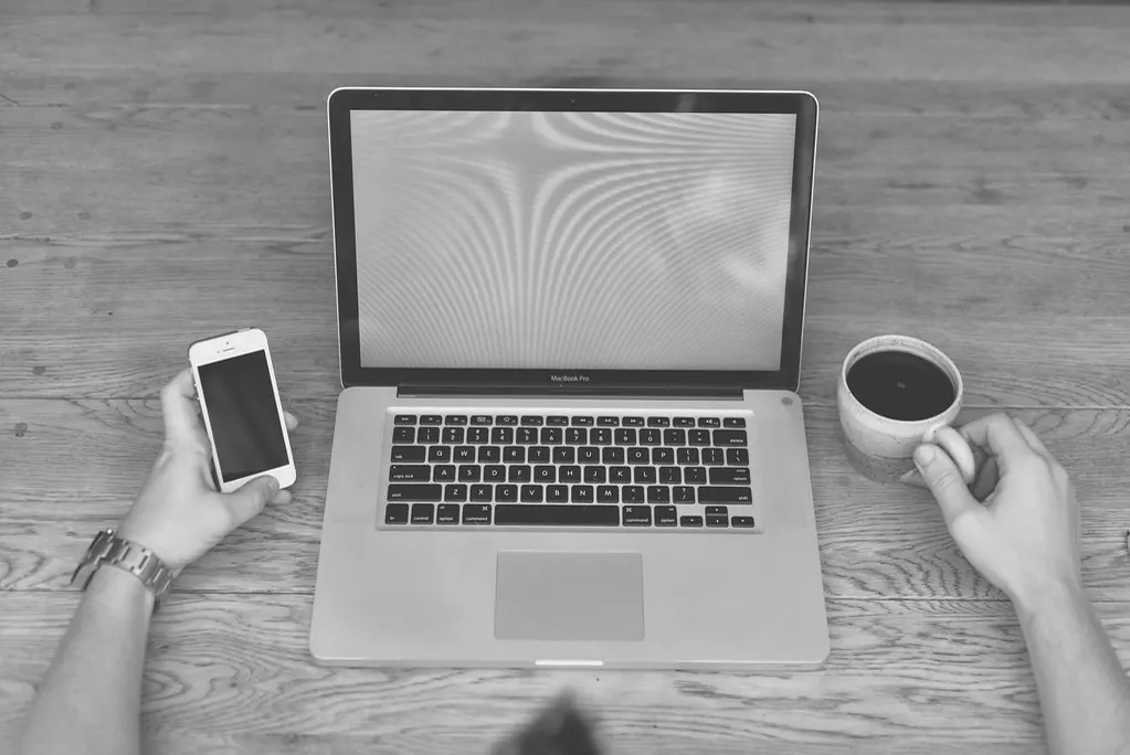 Pessoa com o notebook e o celular sobre a mesa de madeira, ajustando o cadastro de produtos da loja para o comércio agêntico
