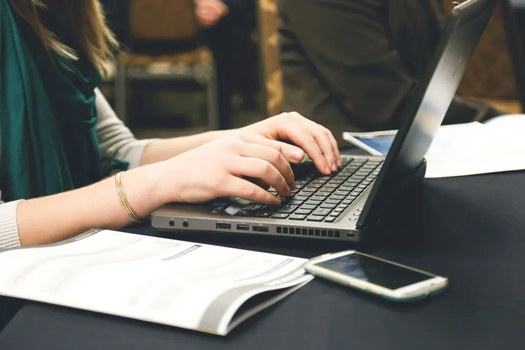 Mulher digitando no notebook com celular e papéis sobre a mesa, pesquisando uma empresa pela busca do ChatGPT