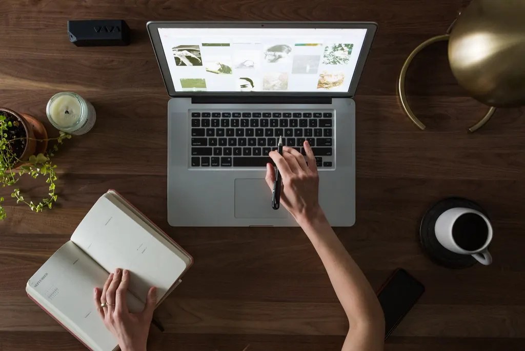 Pessoa trabalhando em um notebook sobre uma mesa de madeira, com agenda aberta e café, organizando tarefas para um agente de IA