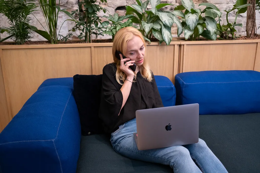 Mulher no sofá falando ao telefone com o notebook no colo, atendendo um contato que chegou pelo anúncio