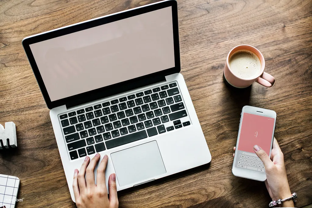 Pessoa usando notebook e celular sobre mesa de madeira com xícara de café, testando IA gratuita para pequenas empresas