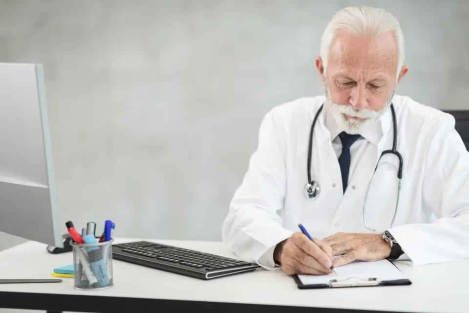 Médico de jaleco escrevendo numa prancheta à mesa do consultório, enquanto a inteligência artificial para clínicas cuida da parte administrativa