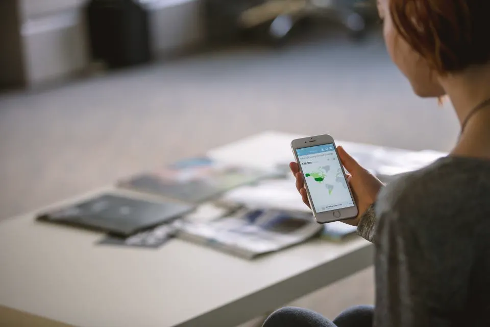 Mulher segurando um celular com um mapa na tela, sentada diante de uma mesa com revistas